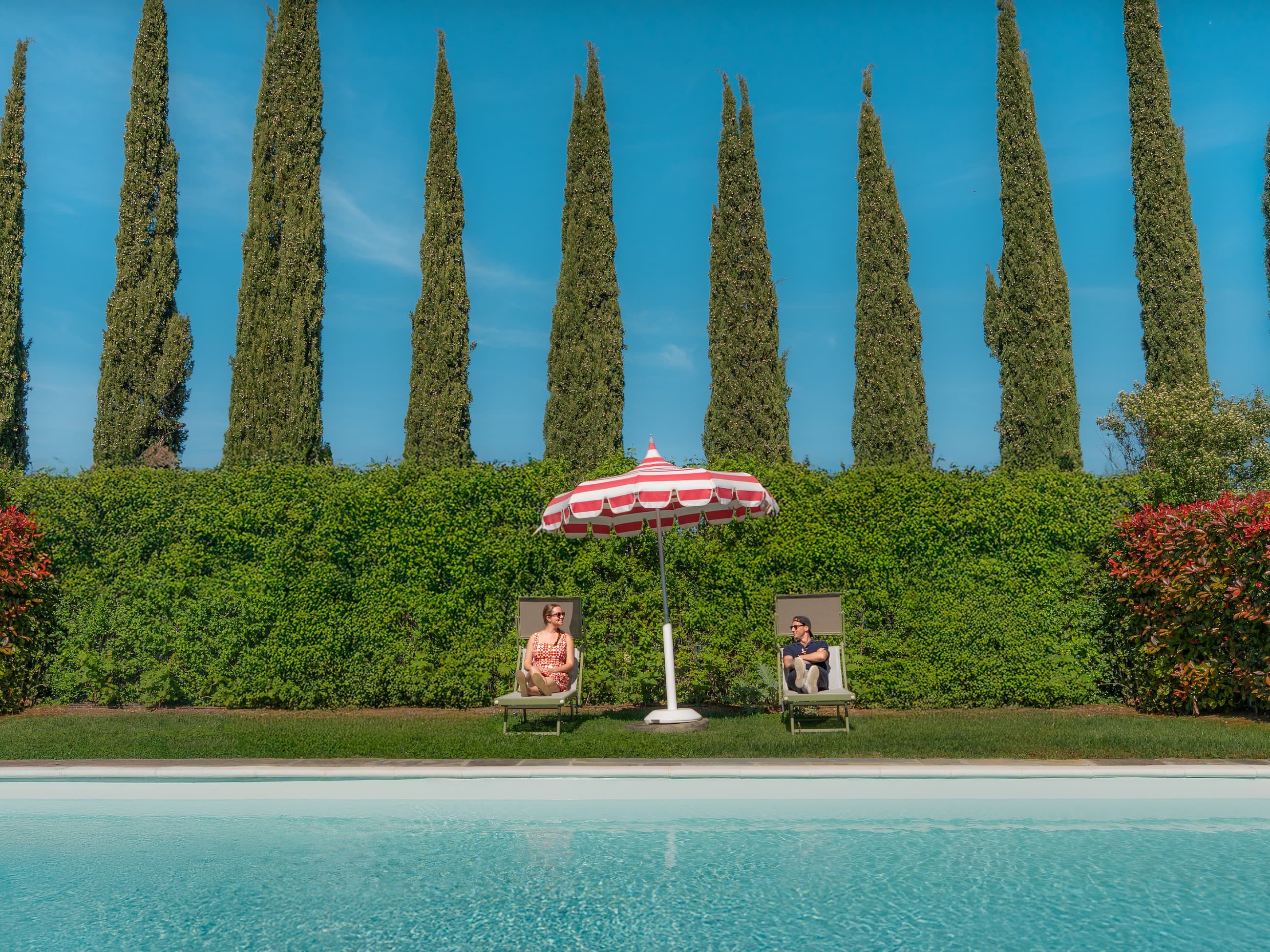 Sam Morgan and Ellie Harwood sitting poolside beneath a striped umbrella, framed by cypress trees in Italy