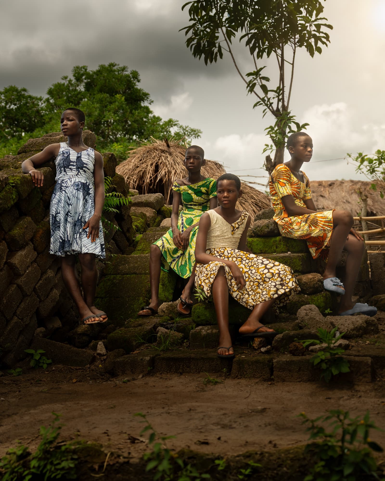 Four girls in patterned dresses seated on stone ruins under a stormy sky, Ghana