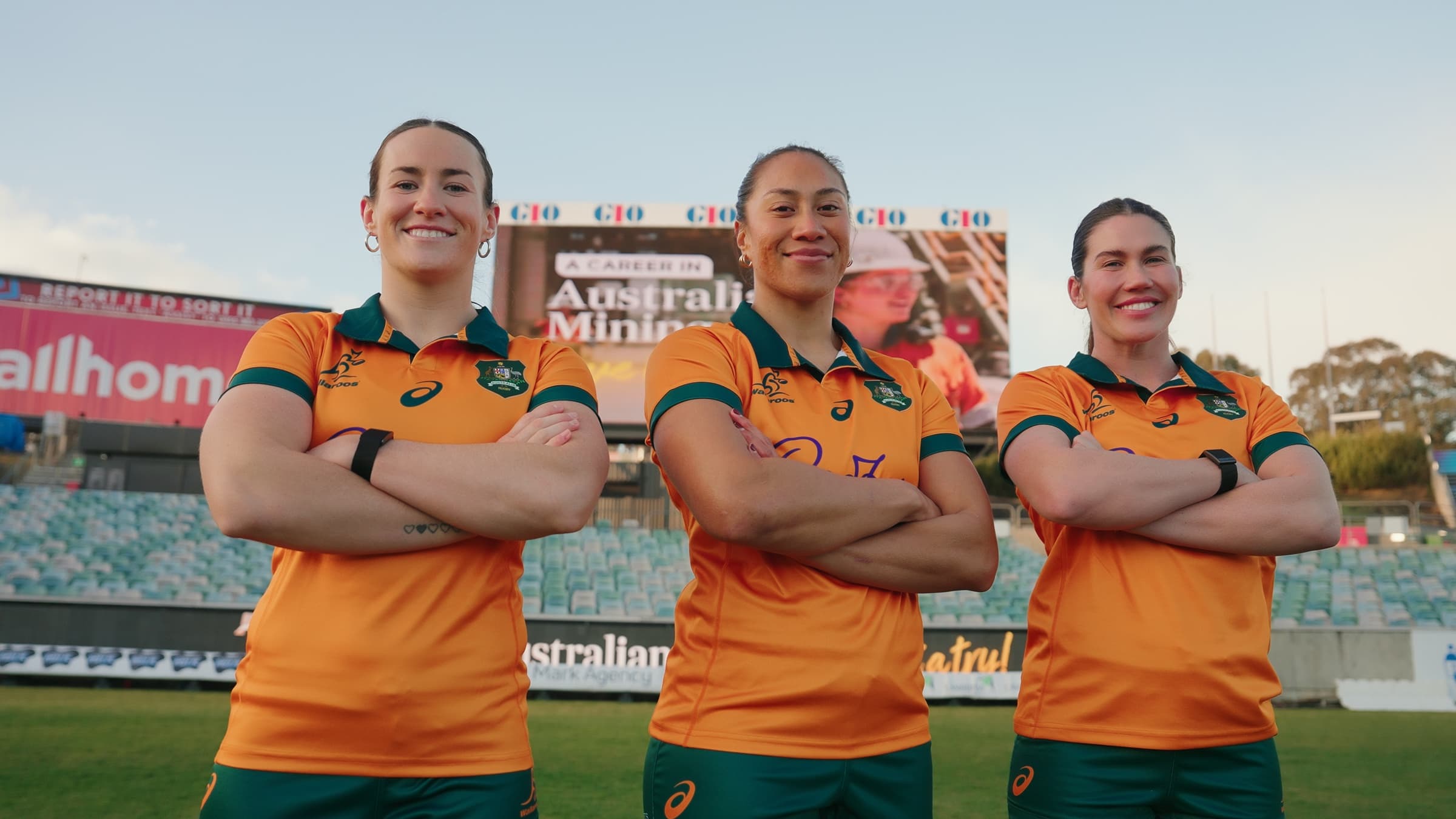 Three Wallaroos players standing arms crossed on the pitch at GIO Stadium, Canberra, in front of the 'A Career in Australian Mining' big screen